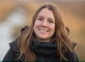 A portrait of a woman with long brown hair and a black jacket standing outdoors. 
